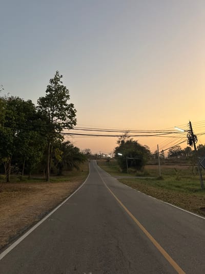 The image depicts an asphalt road ascending uphill and curving slightly left, viewed during early morning or late afternoon in Ban Huai Sai, Thailand. The sky exhibits a gradient from light blue overhead to warm orange and yellow at the horizon. The road features a solid white line on its left edge and a solid yellow center line.
To the left of the road, a strip of dry, reddish-brown earth transitions into dense vegetation, including tall, leafy trees and several palm trees. Overhead, multiple black utility wires stretch horizontally, supported by a wooden utility pole visible on the left.
To the right of the road, the ground is covered in dry grass and sparse green vegetation, bordering what appears to be an open field or agricultural land in the distance. Further along the road on the right, additional utility poles are visible, one featuring an illuminated streetlamp.
In the far distance, indistinct dark figures, possibly two individuals, are visible ascending the road towards the crest of the hill. No immediate interactions or specific actions are discernible among the subjects.