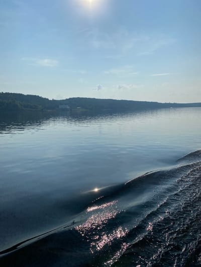 The image depicts a wide body of calm water with gentle ripples, reflecting the bright, hazy sunlight from the upper right corner. A line of trees borders the far side of the water, with a building visible amongst the trees. In the foreground, a wake is forming, with small waves and white foam cresting on the dark water, indicating movement through the water. The sky is a pale blue with scattered wispy clouds and the sun is prominently featured as a bright, blown-out circle. This scene is likely a natural landscape, possibly a lake or a wide river. The mention of Ol'khovskoye, Russia, places the geographical context of this natural scene. There are no people or animals visible in the image.