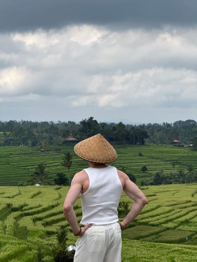 The image shows a person wearing a white tank top, white pants, and a woven conical hat standing in a rice field in Penebel, Indonesia. The person faces away from the camera, looking towards the rice terraces. The terraces are green and stretch out towards the horizon, with trees lining the top of the landscape. The sky above is overcast. The person's arms are bent with their hands on their hips. The rice terraces suggest the location is likely near Jatiluwih.