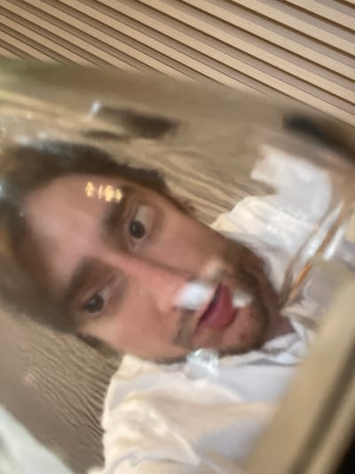 The media file is a close-up, upside-down photograph of a person's face, viewed through a reflective, possibly plastic, surface. The subject appears to be a young adult male with dark hair and fair skin. He is wearing a white garment, possibly a shirt or robe, with some textured details. His eyes are open, and his gaze is directed slightly to the left of the camera. His lips are parted, with a small white mark visible on his upper lip, possibly a reflection or a substance. There are subtle hints of gold or yellow glitter on his cheekbone.

The background consists of diagonal lines, suggesting a textured surface like wood paneling or blinds, and a blurred area of light beige or cream. The lighting is soft, creating reflections on the surface through which the subject is viewed, adding to the abstract and somewhat distorted quality of the image.

There are no visible text elements in the image. The overall impression is intimate and slightly disorienting due to the perspective and the reflective surface. The emotional tone is neutral, with a hint of curiosity or introspection conveyed by the subject's gaze. The location context of Cuautitlán Izcalli, Mexico, does not provide specific visual cues within the image itself.