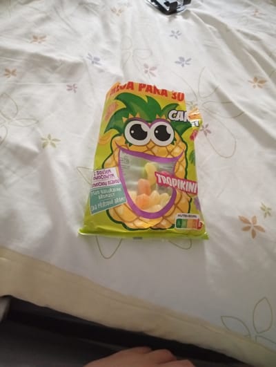 The image shows a bag of Tropikini candies with a pineapple character on it. The bag is yellow and green, and it is lying on a white surface with a floral pattern. The candies inside the bag are colorful and appear to be fruit-flavored. A Nutri-score rating of "D" is visible on the bag. The scene is indoors, and the lighting suggests it might be daytime. There is no visible indication of the city of Galonki, Poland, in the image.