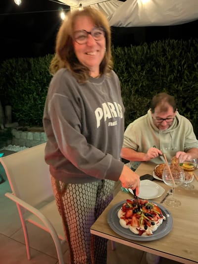 The image shows two people sitting at a table outside, possibly in the evening. A woman in a grey sweatshirt with "PARIS" printed on it is in the foreground, holding a knife and cutting a dessert topped with fruit. A man wearing glasses and a light-colored hooded sweatshirt is seated to her right. He is holding a glass containing liquid and what appears to be crackers. The table also has plates and glasses. In the background, there is greenery and what seems to be an outdoor lighting setup. The location is Veauche, France.
