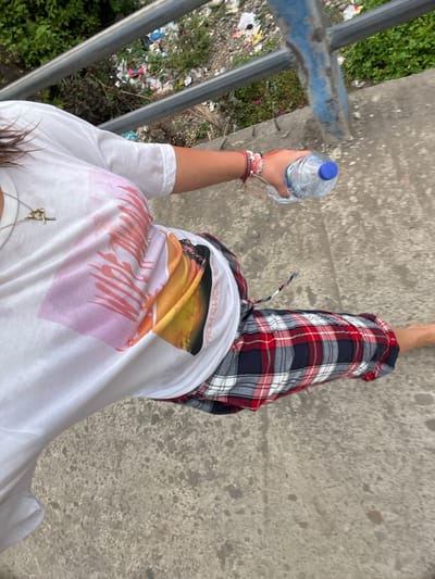 A partial overhead view captures an individual walking forward on a textured grey concrete surface in Desa Batu Layar, Indonesia. The subject, an adult, is visible from the upper torso to the lower right leg, wearing a white short-sleeved t-shirt with a multi-colored graphic design featuring abstract pink/orange elements and a sunset/sunrise scene with "FROM SUNRISE TO" text. Plaid shorts or pants in red, white, and dark blue patterns are worn. A silver-colored necklace with a cross pendant is on the subject's neck, and multiple multi-colored bracelets adorn the right wrist. The right hand holds a clear plastic water bottle with a blue cap, containing clear liquid. The subject's right foot, visible at the bottom right, is bare. A two-bar metal railing, colored grey and blue, runs horizontally across the upper background, separating the walking surface from a lower area. This lower area is characterized by dense green vegetation interspersed with a substantial accumulation of discarded refuse, including plastic bags and various debris.