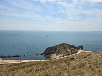 The image shows a wide, high-angle view of the coastline near West Lulworth, United Kingdom. In the foreground, dry, golden grasses cover a sloping hillside. A dirt path winds down the hill towards the sea. Further down, a sandy beach is visible with several small groups of people walking or standing.
The sea occupies the middle ground of the image, a deep blue color with sunlight glinting off its surface. In the distance, a lone sailboat is visible on the horizon.
The most prominent feature is a large rock formation jutting out into the sea, characterized by a natural archway. This iconic landmark, Durdle Door, is covered in green vegetation. Many people are gathered on the paths and near the archway, exploring the area. The sky above is a pale blue with streaks of white clouds. The overall scene is one of a sunny, popular tourist destination on the coast.