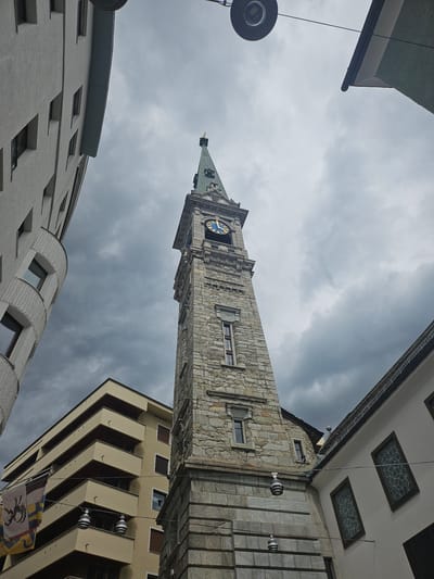 This is a low-angle, wide-shot photograph taken in the city of St. Moritz, Switzerland. The image features a tall, stone clock tower with a green, pointed roof and a golden finial at its apex. A clock face with blue numerals is visible on the tower. To the left of the clock tower, a modern, white building with multiple windows curves away from the viewer. To the right, a white building with dark-framed windows, one of which has a patterned glass, is partially visible. In the background, a yellow building with stacked balconies and dark awnings is seen. A flag with a yellow and blue design and an emblem of a winged creature hangs from a wire. Two decorative, silver outdoor lights are suspended from wires in the foreground. The sky is overcast with dark, heavy clouds. The scene appears to be a street view within the urban environment of St. Moritz.