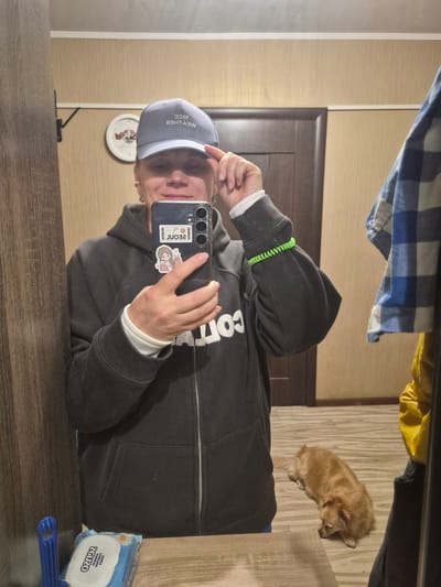 A medium shot captures a person in a grey hooded jacket and a blue baseball cap, taking a mirror selfie. The person's right hand adjusts the cap, while their left hand holds a smartphone. The phone displays two stickers: one with Korean text that reads "SEOUL" and another of a cartoon girl. The background reveals a room with wooden doors and walls. A wall clock with Roman numerals is visible on the left. A small, light brown dog lies on the wooden floor to the right of the person. A package of wet wipes and a blue object are on a table in the foreground. The overall setting appears to be domestic. The image does not provide any specific information that would allow it to be located in the city of Nyagan, Russia.