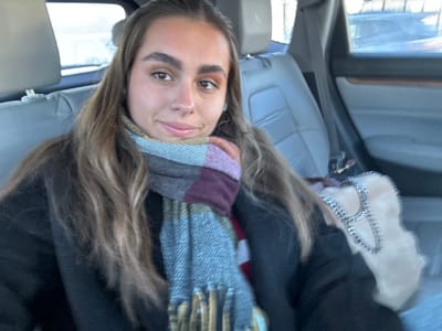 A selfie of a young woman in a vehicle. She wears a dark coat and a patterned scarf with fringe. Her hair is long and light brown. A fuzzy white bag with a stitched "A" is visible. The interior of the car has light gray seats. Outside the window, a blurred view hints at a street or parking lot. The image is rotated, making the woman appear upside down.