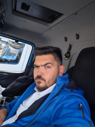 The image, taken in Kolbermoor, Germany, features a man inside what appears to be a vehicle cabin, likely a truck. The man, positioned with his head on the left and body extending to the right due to the image orientation, has dark hair, a beard, and a mustache. He wears a vibrant royal blue jacket with black zipper details, layered over a white collared shirt and a black garment underneath. His gaze is directed towards the camera with a neutral expression.
The interior of the vehicle is characterized by grey, textured surfaces. A window to the man's right reveals a clear reflection of another blue vehicle, possibly another truck, with a chrome mirror casing visible. This suggests an outdoor setting, perhaps a parking lot or industrial area, during daylight hours.
On the grey surface next to the man's head, a black tree-shaped air freshener is visible. Text on the air freshener clearly reads "WUNDER-BAUM" and "Made in EU". Several small black clips or hooks are also present on the surrounding interior surfaces. The man appears to be leaning back or in a semi-reclined position. The overall impression is a casual moment captured within a commercial vehicle.