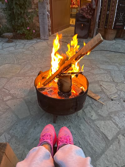 The image captures a warm outdoor scene focused on a crackling bonfire in a metal fire pit. The ground is paved with large, irregular flagstones. In the foreground, a person's legs are visible, wearing bright pink sneakers and light-colored pants, suggesting they are seated and enjoying the fire.

The fire pit is filled with burning logs and embers, with flames leaping upwards. A piece of wood extends out of the fire pit, angled towards the viewer. In the background, to the left, is a stone wall covered in greenery and some pink roses, with a rustic wooden sign that appears to be a menu. Further back, to the right, is a dark-colored speaker and some decorative woven textiles hanging on a wall, with a wicker basket nearby. The overall ambiance is cozy and inviting.

The image was taken in Gusinje, Montenegro, a town known for its natural beauty and traditional architecture. The setting appears to be an outdoor area, possibly a courtyard or patio of a restaurant or guesthouse, given the presence of a menu and the rustic decor. The lighting suggests it is either dusk or early evening.