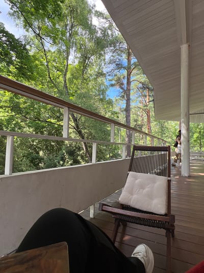 The image depicts a partially occupied outdoor seating area adjacent to a forested environment, likely located in or near Vnukovo, Russia. In the foreground, a person's legs, clad in black pants and white sneakers, are visible, suggesting they are seated. A wooden chair with a white cushion is positioned centrally, facing away from the camera. The chair itself is made of dark wood with a woven seat and back. Beyond the seating area, a balcony or deck with wooden flooring extends. A white railing runs along the edge of the balcony, separating it from the dense green foliage of trees. The trees appear to be a mix of deciduous and coniferous species, with sunlight filtering through their leaves. In the background, a portion of a building's exterior is visible, including a white pillar and a white, sloped roof. Two individuals, one with dark hair and wearing a white top and beige pants, and another partially obscured figure, are standing further down the deck, engaged in an interaction. The overall impression is one of a relaxed, natural setting.