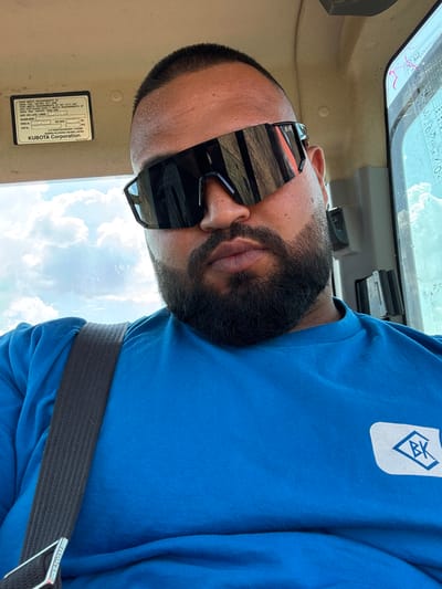 A man with a beard and sunglasses is inside a vehicle. He is wearing a blue t-shirt with a logo on the left side of his chest. A seatbelt is visible across his chest. The man appears to be in an agricultural setting, with the sky and clouds visible through the vehicle's windows. A label with text and a "Kubota Corporation" logo is affixed to the vehicle's interior. The man is not actively engaged in any discernible action. The location is identified as Troyanovo, Bulgaria.