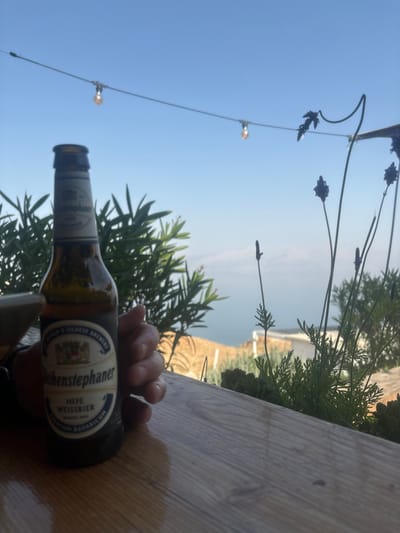 The image depicts an outdoor daytime scene within the Megilot Yam HaMelakh Regional Council, Palestinian Territory. A light-brown wooden tabletop, exhibiting a visible grain, occupies the lower foreground. An adult's right hand, with light skin tone and visible knuckles, is positioned on the left, holding a brown glass bottle of "Weihenstephaner Hefe Weissbier." The bottle features a white and blue label adorned with a crest and text, and a gold-colored bottle cap is partially visible.
In the midground, directly behind the hand and bottle, dense green foliage is visible. To the right, several slender lavender plants with dark purple, spike-like flowers rise from an unseen planter or garden bed. Above the vegetation, a string of outdoor lights, featuring multiple illuminated, warm-toned bulbs, extends horizontally across the frame.
The background reveals a large, calm body of water, light blue-grey in color, stretching to the horizon. This body of water is consistent with the Dead Sea. Beyond the water, faint, light-brown landforms (hills or cliffs) are discernible in the distance. The sky above is clear and uniformly blue, indicating bright, sunny environmental conditions.
