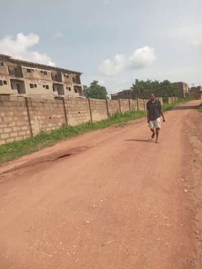 The image shows a man walking along a red dirt road in what appears to be the Achara Ndiagu Omegio area of Nigeria, as evidenced by the distinct reddish soil common in the region and building styles. The man is wearing a dark shirt and light-colored shorts with sandals. Beside the road, a concrete block structure under construction is visible, suggesting an ongoing development in the area. The sky above is partly cloudy. The environment is dry and dusty.