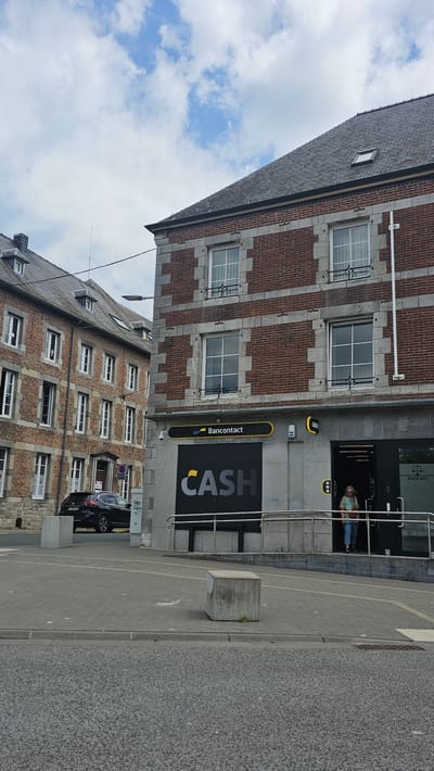 The image depicts a street view in Philippeville, Belgium. On the right side of the frame is a brick building with stone accents, housing a "CASH" establishment. A yellow sign with "Bancontact" is visible above the entrance. A woman is seen exiting the "CASH" establishment. To the left of this building, another brick building with multiple windows is partially visible, suggesting an urban or semi-urban environment. A dark-colored car is parked on the street in front of the buildings. The ground is paved with interlocking stones and asphalt. The sky is partly cloudy.