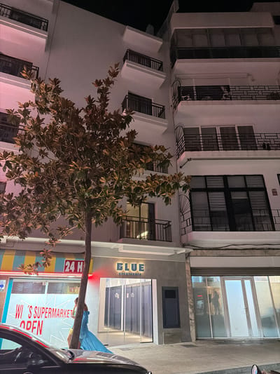 This is a nighttime, eye-level exterior shot of a multi-story building in Sant Antoni De Portmany, Spain. The building is illuminated with pink and white lights, creating a soft glow. A tree with large, leathery leaves stands in front of the building on the left side of the frame, its trunk crossing the foreground. To the right of the tree, a storefront with the name "BLUE APARTMENTS" is visible. Further left, another storefront is illuminated with bright yellow and blue striped signage, displaying "24 H" and "WI'S SUPERMARKET OPEN" in large red lettering. A person in a bright blue dress is partially visible behind the tree, facing away from the camera. A black car is parked in the foreground, partially obscuring the lower part of the scene. The overall impression is that of a street-level view of commercial and residential buildings at night.