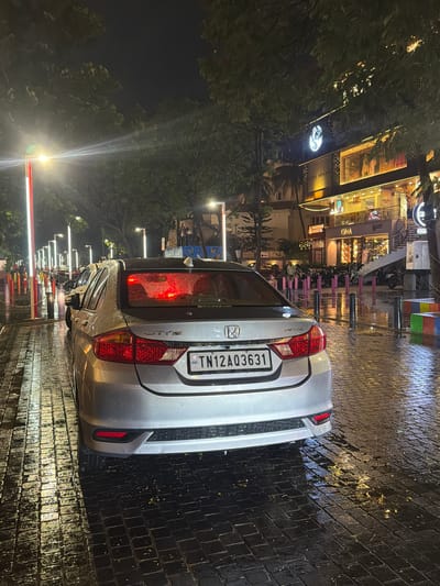 The image depicts a rear view of a silver Honda City sedan parked on a wet, cobblestone street at night. The car's brake lights are illuminated red, reflecting on the wet surface. A license plate with "TN12 A03631" is visible. To the left, a line of illuminated poles with red and white markings extends into the distance, indicating a pedestrian walkway or street demarcation. Trees line the street, and beyond them, a building with multiple illuminated floors is visible. The building's signage includes the words "OMA" and "Filli cafe". The scene suggests a commercial area in Chennai, India, possibly in the evening after rain. The lighting is atmospheric, with streetlights and building lights creating reflections on the wet ground. No people are clearly visible in the immediate vicinity of the car, but some figures can be faintly seen in the background near the building.