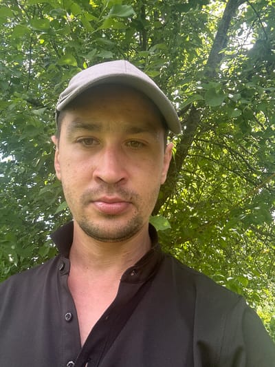 This is a close-up, low-angle selfie of a man taken outdoors. The man is wearing a light grey flat cap and a black collared shirt. His face is turned towards the camera, and he has light brown eyes, a light beard, and fair skin. He is looking slightly to his left.
The background is dominated by lush green foliage, suggesting a natural outdoor setting like a park or garden, likely during daylight hours given the bright lighting. The way the leaves and branches fill the frame implies the photographer is situated beneath trees. The orientation of the image is rotated 90 degrees counter-clockwise from standard portrait orientation, with the man's head appearing at the bottom of the frame.
The context provided indicates the location is Yelmyachi, Russia. No specific activity is depicted, but the casual attire and outdoor setting suggest a relaxed moment. There is no visible text in the image. The overall mood is informal and natural.