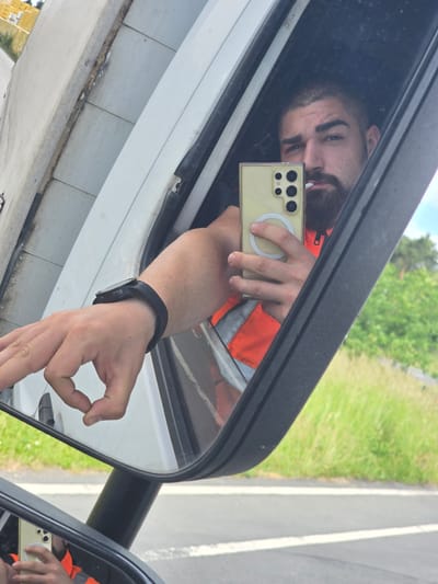 An eye-level selfie captures a man's reflection in a vehicle's side mirror, presenting a distorted and rotated perspective. The man, with a beard and dark hair, looks directly towards the camera of his smartphone, which he holds up with his right hand. The phone is yellow or gold, encased in a clear cover with a prominent circular MagSafe-like ring on the back, and multiple camera lenses are visible. He is wearing an orange reflective safety vest, indicative of outdoor or roadside work.

His left arm, adorned with a black smartwatch, extends out, and his hand forms an "OK" gesture. The vehicle's white exterior and part of its structure are visible around the mirror. The background, seen in the mirror's reflection, depicts a bright daytime scene of a road with a white line separating it from a stretch of green grass and distant trees under a clear sky. This suggests a location beside a rural or suburban road. The image is taken in Laußig, Germany. No discernible text is visible in the image.