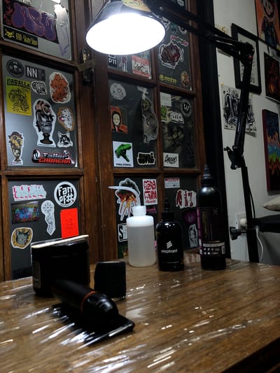 This image shows a close-up, high-angle view of a tattoo artist's workspace. The scene is illuminated by a bright adjustable lamp with a large circular light source. Behind the lamp is a wooden cabinet or display case covered with various stickers. The stickers display a range of images, including cartoons, logos, graffiti art, and text. Several bottles of tattoo-related products, such as ink and stencils, are arranged on a wooden surface covered in plastic wrap. The brands "Wadron" and "Elephant" are visible on the product labels. No people are visible in the image. The overall impression is that the image was taken in a tattoo studio. The location of the scene is not provided.