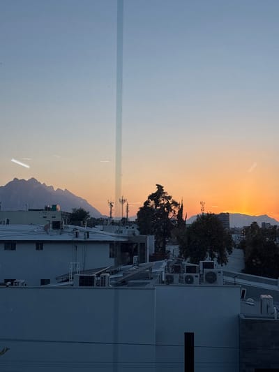 The image shows a cityscape in San Pedro Garza García, Mexico, at sunset. Mountains are visible in the distance, with orange and yellow light from the setting sun. The foreground is dominated by buildings with numerous air conditioning units on the rooftops. There are trees interspersed among the buildings and telecommunications infrastructure visible. The sky ranges from light blue to orange with the setting sun. The image seems to have been taken through a window as evidenced by a reflection or streak on the left side.