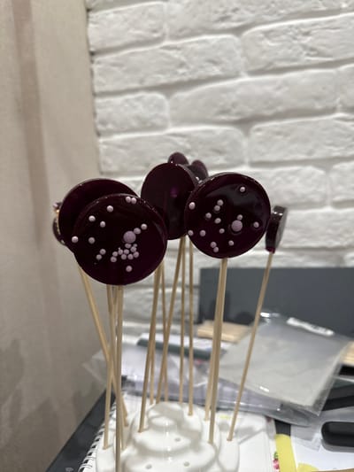 A close-up, eye-level shot captures a cluster of round, dark purple lollipops arranged on wooden sticks. The lollipops are decorated with small, light pink spherical candies, some clustered together in patterns and others scattered individually across the translucent surfaces. They are held upright in a white, multi-holed display stand, which sits on a dark surface, possibly a table. In the background, a white brick wall provides a textured backdrop. The lighting is moderately bright, with some soft shadows and reflections on the lollipops, indicating an indoor setting. The overall impression is of a confectionary display. There is no information in the image to suggest a specific location in Ulan-Ude, Russia.