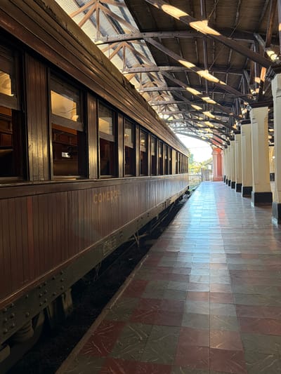 The image shows a view of an old train car at a station platform in Asunción, Paraguay. The train car is made of dark wood and has several windows. The platform is paved with checkered tiles in shades of red and grey. An open-sided roof structure, supported by thick wooden beams and corrugated metal sheeting, covers the platform. Sunlight streams through the gaps in the roof, casting patterned shadows on the platform and train car. The train car has the word "COMEDOR" stenciled in yellow on its side. The overall scene suggests a historic or preserved train station.