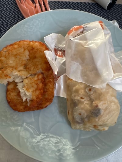This overhead shot shows a light blue plate with two food items on it, positioned on a dark blue and white chevron-patterned placemat. To the right, there is a light beige burrito, partially wrapped in white paper. The burrito appears to be filled with dark ingredients, possibly beans and meat, and the tortilla shows signs of being grilled. To the left of the burrito is a golden-brown hash brown. It has a crispy exterior and a fluffy, shredded interior, with a bite taken out of it, revealing the white potato shreds. The plate has a subtle embossed pattern of wavy lines. In the background, to the left of the plate, a corner of a white and black object, possibly a camera lens cap or a small container, is visible. A portion of a pale pink utensil, with a ridged surface, rests on the placemat to the left of the plate. Faint orange and black text, likely from a brand logo, is visible on the white paper wrapping the food items, but it is largely obscured. The lighting suggests it is daytime, possibly indoors given the placemat and plate. There are no people visible in the frame.