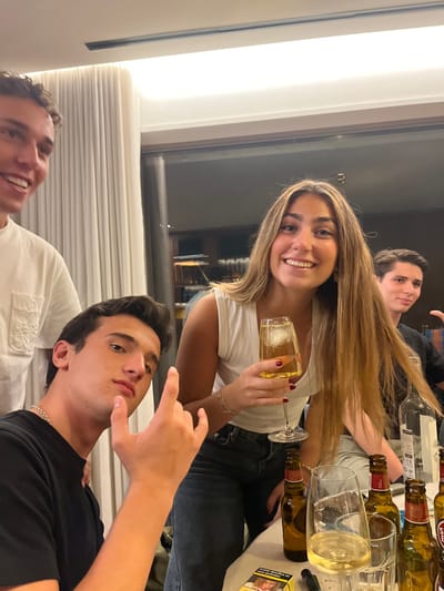 This image depicts an indoor social gathering involving four young adults at a residence in Lordelo do Ouro e Massarelos, Portugal.
A female with long, light brown hair, wearing a white v-neck top and dark jeans, smiles directly at the camera. She holds a stemmed glass containing an amber liquid with ice in her right hand; her fingernails are painted dark red. To her left, a male with dark hair, wearing a black t-shirt and a silver chain necklace, looks at the camera while making a "devil horns" hand gesture with his right hand. Partially visible on the far left, another male with light brown curly hair, wearing a white t-shirt, smiles towards the left. On the right, a fourth male, wearing a dark top, is partially visible, smiling towards the right.
The group is gathered around a white table laden with beverages. Visible items include several empty and partially filled amber glass bottles, identifiable as Super Bock beer. A clear glass bottle, likely containing spirits, and a wine glass with a light-colored liquid are also present. A yellow and white Marlboro cigarette pack and a small black cylindrical object, possibly an e-cigarette, are on the table surface. The background features a large window or glass door with light-colored curtains to its left, revealing a dimly lit interior or exterior space beyond. Recessed lighting fixtures are visible on the ceiling.