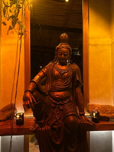 A wooden Buddha statue is the main subject of the image. The statue is situated indoors, possibly in a room or a building in Pererenan, Indonesia. The setting is illuminated with a warm, perhaps candlelit glow. The Buddha is positioned between two wooden frames, which may be part of a window or a room divider. There is a dark reflection on the glass, suggesting it is nighttime outside. A plant is visible in the bottom left corner. Two small, dark containers are also in the frame, suggesting possibly candles.