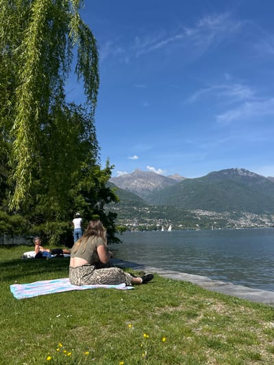 The photograph shows a scenic view of Locarno, Switzerland, featuring a person sitting on the grass near a body of water. The individual is lying on a striped towel on a grassy bank, near the edge of the lake. In the background, there is a clear blue sky with scattered clouds, a view of the lake, and mountains. There is a city partially visible, nestled along the shoreline and up the mountainside. A weeping willow tree is visible in the foreground on the left side of the frame. Other people are in the background.