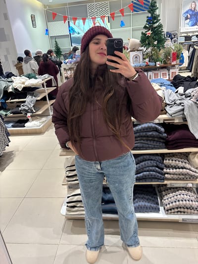 A woman wearing a maroon puffer jacket and jeans stands indoors, likely in a clothing store. She is taking a selfie with a black phone, wearing a maroon beanie. Clothes racks displaying folded garments surround her. There's a Christmas tree decorated with lights and ornaments visible in the background. The floor is tiled and light-colored.