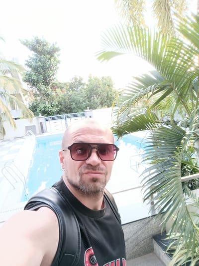 A bald man wearing sunglasses and a black sleeveless shirt is taking a selfie by a swimming pool. He has a goatee and is wearing a hoop earring in his left ear. To the left of the man is a swimming pool with ladders and to the right are palm trees. The man appears to be in Anajpur, India.