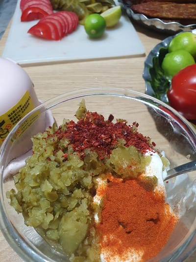 The image depicts a close-up view of a glass bowl containing a mixture of chopped green pickles, a dollop of white cream or yogurt, and a generous sprinkle of red chili flakes and paprika. A fork is partially visible, resting in the mixture. In the background, sliced tomatoes, limes, and a whole tomato are arranged on a white cutting board. Further in the background, a metal dish holds what appears to be grilled meat. A pink electric device with a yellow label containing Chinese text is also visible to the left. The scene is set on a wooden table. The image does not contain any information that would allow for its location to be specified in Kheyrabad, Iran.