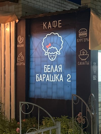 The image shows the facade of a cafe named "БЕЛАЯ БАРАШКА 2" (White Lamb 2). The sign features a stylized white sheep with a red bow on its head, and text in white neon lights. To the left of the sheep, there are icons and text for "МИДИИ" (Mussels) and "ДЕСЕРТЫ" (Desserts). To the right, there are icons and text for "БУРГЕРЫ" (Burgers) and "САЛАТЫ" (Salads). A small icon with a sheep silhouette and the number "662" is visible on the far right. The cafe appears to be located in a city, with a brick wall and awning visible in the background. A decorative metal fence with some greenery and flowers is in the foreground. The overall impression is of an outdoor seating area for a casual dining establishment. The city of Shchelkino in Ukraine is not directly identifiable from the image itself, but is provided as context.