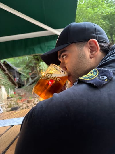 A man wearing a black baseball cap and a dark jacket is drinking from a glass of amber-colored beer. The glass has a logo that reads "Nikšićko 130 godina". The man is seated at a wooden table, possibly outdoors. In the background, there is greenery and a rustic-looking structure, suggesting a natural or semi-outdoor setting. The scene does not provide any specific visual information that can be used to pinpoint a location within the city of Brijeg, Montenegro.