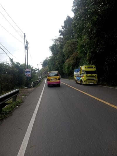The image captures a view from a moving vehicle on a two-lane asphalt road, likely in Indonesia, given the visible text. The sky is overcast, suggesting a cloudy day with diffused lighting.
In the foreground and midground, two vehicles are traveling away from the viewer. In the left lane, closer to the camera, is a yellow minivan with a distinct pink rear bumper. Further ahead in the right lane is a bright yellow truck, possibly a small delivery truck, with a blue tarp covering its cargo bed. The truck has "SCANIA" written on its front grille and some blue text, which appears to be "NGANTER", visible on its side.
The road itself has clear markings: a solid white line separates the left shoulder from the main driving lane, and a double solid yellow line divides the two lanes of traffic.
On the left side of the road, there is a paved shoulder or narrow sidewalk bordered by a metal guardrail. Behind the guardrail, dense green foliage and bushes are visible. Further along the left side, utility poles with numerous overhead wires extend into the distance. Attached to one of these poles is a blue and white sign featuring the "Indomaret" logo (an Indonesian convenience store chain) and the text "300 M" below it, indicating a store location 300 meters ahead. Below this text are small images of various products.
The right side of the road is bordered by a steep embankment or hillside covered in dense, dark green trees and lush vegetation, forming a natural wall along the road. The overall impression is of a road winding through a natural, possibly mountainous or hilly, landscape.