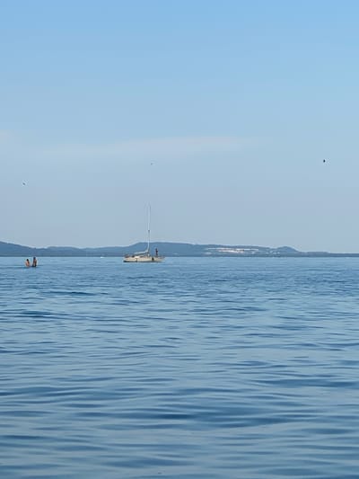 The image depicts a serene scene on a body of water, likely Lake Neuchâtel in Milvignes, Switzerland. The composition is dominated by the calm, blue water with gentle ripples, reflecting the clear sky. In the background, a hazy silhouette of distant hills suggests a tranquil landscape.
Several subjects are visible. On the water, a white sailboat with a tall mast is anchored. A few people are present on the boat, and a couple more are seen in the water near the bottom of the frame, seemingly enjoying the day. The presence of a few birds in flight adds to the natural ambiance.
The time of day appears to be late afternoon or early evening, given the soft, diffused light and the clear, pale blue sky with no harsh shadows. The weather seems pleasant and calm, ideal for a day spent on the water. The overall mood conveyed is one of peace and relaxation. There is no visible text in the image.