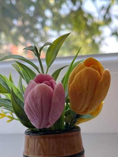 The image shows a close-up of a small, artificial floral arrangement in a brown and black striped vase. The arrangement features two prominent tulip-shaped flowers, one pink and one orange, surrounded by green artificial leaves and a few small yellow accent flowers. The background is blurred, suggesting an outdoor setting with bokeh effects, possibly a window with sunlight filtering through trees. The scene is likely located indoors, near a window, in the city of Branipole, Bulgaria. There are no people or animals visible in the image. The lighting appears to be natural daylight.