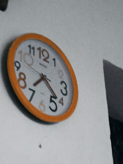 The image shows a close-up, slightly distorted view of an orange-rimmed analog clock mounted on a white wall. The clock face displays black Arabic numerals, with some of the numbers appearing slightly faded or a different color, possibly due to lighting or age. The hour hand is pointing between the 7 and the 8, appearing closer to the 7. The minute hand is pointing directly at the 5. The second hand is not clearly visible. The clock's hands are a dark brown color. The background is a plain, textured white wall. To the right of the clock, a dark, angled shadow is cast on the wall, suggesting a light source is present but not within the frame. There is a small dark speck on the wall below the clock. No people or any other objects are visible in the frame. The image quality is grainy, indicating it was taken with a low-resolution camera or in poor lighting conditions. The scene is indoors. There is no information or visual cues to suggest the location is in Ceyranbatan, Azerbaijan.