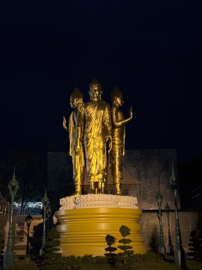 The image shows a nighttime scene featuring several golden Buddha statues. The statues appear to be standing on a large, fluted golden base. One statue is in the foreground, facing left, with its arm raised. Behind it, two more statues are visible, also facing left. The statues are illuminated by artificial light, casting a warm glow against the dark night sky.
In the background, there are tall, ornate lampposts with a dark green color. Behind the lampposts, a large, textured stone wall is visible, possibly part of a building or monument. There are also trees and foliage present, suggesting an outdoor setting. The ground appears to be covered in grass on the right side of the image.
The overall impression is one of solemnity and reverence, typical of religious sites. The golden statues and the nighttime setting create a dramatic and striking visual. The reflections of the lampposts in what appears to be a wet surface or water add another layer of visual interest to the scene.
There is no visible text in the image. The time of day is clearly night due to the dark sky and artificial lighting. The weather appears clear, as there are no indications of rain or fog. The location is likely a temple or a public monument in Phnom Penh, Cambodia, given the context provided.