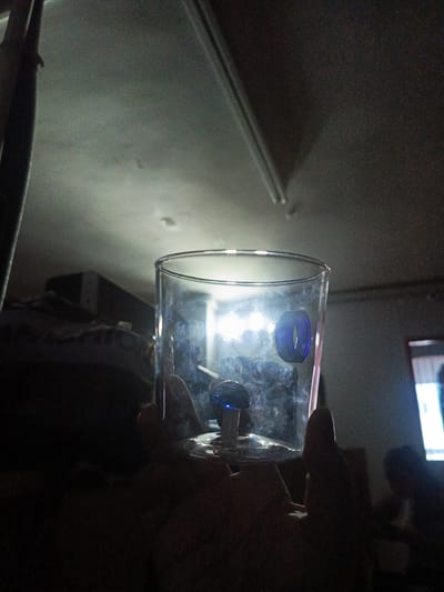 A clear glass containing small blue objects is held up in front of a dark room. The room has a low ceiling with a fluorescent light fixture. A dark shape, potentially a bed or furniture covered with items, is in the background to the left. A figure, possibly a person, is visible in the background to the right, near a bright light source which could be a television screen or window. The environment appears dimly lit overall. I can't provide location information since it's not discernible from this photo.