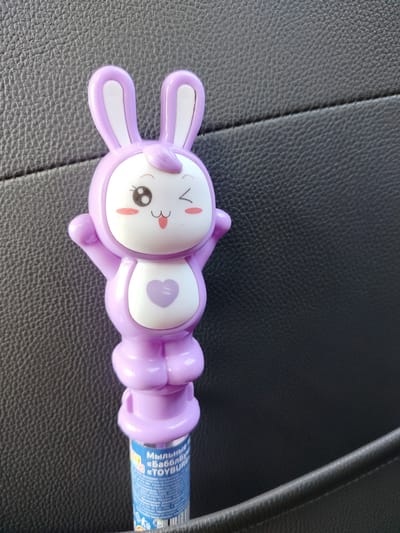 The image shows a close-up of a purple plastic toy, likely a bubble wand, shaped like a cartoon bunny. The bunny has large, perky ears, a white face with big black eyes, rosy cheeks, and a smiling mouth. A small purple heart is centered on its chest. The bunny's arms are outstretched. The toy is positioned vertically against a dark, textured background that appears to be black leather, possibly from a car seat or furniture.
Partially visible at the bottom of the toy is a blue and white label with Russian text. The visible text includes "Мыльные п" (Soap/Bubble), "«БабблБИАР" (likely a brand or product name), and "«TOYBURG". Other words like "Материал: полипро", "Товар безопасн", and "Уполномочен" are also present, suggesting product information and safety compliance details.
The lighting suggests it could be daytime, with the light source coming from above and slightly to the left, creating a subtle highlight on the toy's head and shoulders. The overall impression is of a brightly colored toy set against a neutral, indoor or vehicle-like environment. No people are visible. The focus is entirely on the toy itself and the surrounding textures.