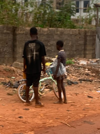 The image, set in Aguleri, Nigeria, captures two young males standing alongside bicycles on a reddish-brown dirt path. The ground in the middle distance contains scattered debris and dry organic matter. In the background, a low, rough-hewn stone wall spans the width of the frame, partially obscured by green foliage, trees, and parts of a white building with visible windows to the far left.

The individual in the foreground is seen from behind, wearing a black t-shirt with a prominent white graphic on the back. This graphic features a stylized cross design above some indistinct, blocky white text that appears to be "919" or similar characters. They also wear dark shorts. Next to this person is a bicycle with a visible yellow or gold-colored frame and white tires.

Slightly behind and to the right, the second individual also stands facing away from the viewer. This person wears a darker, possibly grey or faded purple t-shirt and light-colored shorts or trousers. They are positioned beside a light green or mint-colored bicycle, which also features white tires and yellow accents on its wheels and frame. Both individuals appear to be barefoot or wearing minimal footwear. The scene is illuminated by diffused daylight, suggesting either an overcast sky or indirect sunlight. The two seem to be casually standing, possibly resting with their bicycles. No specific emotions are discernible from their posture or visible features.