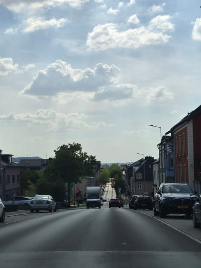 The image shows a street scene in Käerjeng, Luxembourg, on a cloudy day. The perspective is from the driver's seat of a car, looking forward and slightly to the left.
On the right side of the frame is a paved road with a white line marking the edge. Several cars are parked along the side of the road and are also visible driving on the road. The car in the foreground on the right is a white sedan with a license plate that is partially visible. Further down the road, a black SUV with a license plate that reads "TM 0812" is parked.
On the left side of the frame, there are buildings with various architectural styles. Some of these buildings are multi-story residential or commercial properties. Trees are also visible, adding greenery to the urban landscape. In the distance, there appear to be more buildings and possibly hills.
The sky is filled with white and gray clouds, suggesting a partly cloudy or overcast day. There are no people clearly visible in the image. The overall atmosphere is one of a typical European street scene. A traffic light is visible in the distance, showing a red light, indicating that the cars are stopped or slowing down.
The time of day appears to be daytime, likely afternoon, given the brightness and cloud cover.
