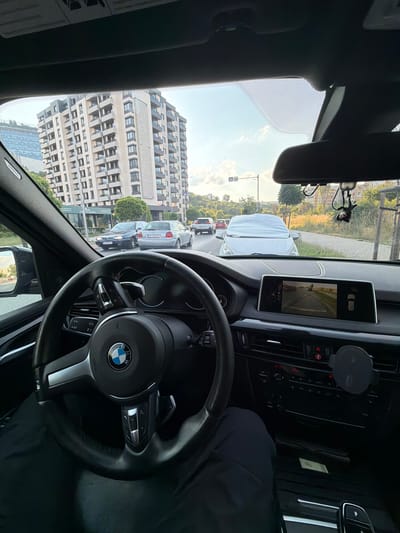 This media file captures an interior view from a car parked on a street in Busmantsi, Bulgaria. The perspective is from the driver's seat, looking forward and slightly to the left. The dominant object in the foreground is the car's black leather steering wheel, featuring a BMW logo in the center. To the right of the steering wheel, the dashboard is visible, including a digital display screen showing a rearview camera feed.

Outside the car, the scene depicts a street with several vehicles lined up, suggesting traffic or a parked situation. A distinctive, modern building with a grid-like facade stands prominently on the left side of the frame, indicating an urban or suburban setting. Trees and greenery are also visible, hinting at a pleasant day.

The sky is bright and appears to be daytime, with some clouds. The overall impression is that of a typical day, possibly during daylight hours, with the car stationary on a street. No people are clearly visible within the car, though a person's shoulder and arm are seen in the dark interior on the right side of the frame. The image is oriented sideways. No discernible text is visible on the steering wheel or dashboard.