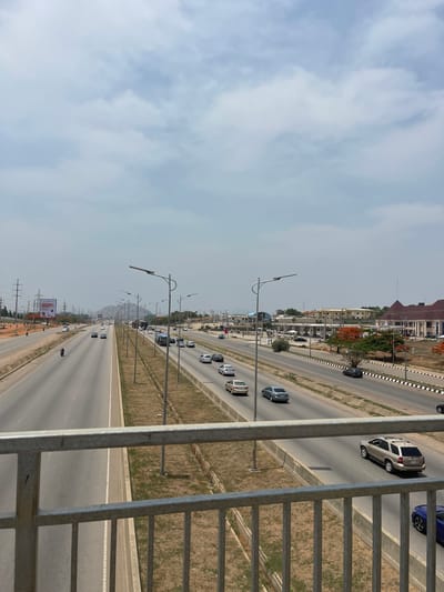 The image depicts a multilane highway in Kuchingoro, Nigeria, viewed from an elevated position, likely a pedestrian bridge. The sky is overcast with scattered clouds. Two sets of lanes run parallel, divided by a median. Cars are visible in both directions. Streetlights line the highway. Buildings and vegetation are visible on the far side of the highway. A metal railing is in the foreground.