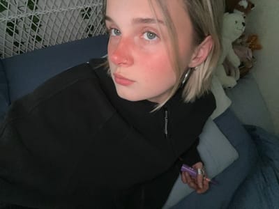 A person is reclining on a blue couch with their head near a pile of stuffed animals. The person is wearing a black hooded sweatshirt and is holding a purple vape pen. They have a nose ring, pale skin, and blonde hair. The background includes a white wire lattice pattern. The location of Whistler, Canada cannot be determined from the image.
