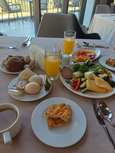 The image depicts a breakfast spread laid out on a table, likely in a hotel or restaurant. The setting appears to be indoors, with a large window in the background showing an outdoor area with chairs and tables, possibly a patio or balcony overlooking a landscape.
On the table, there are several plates and glasses. One plate holds a variety of items: a round bread roll, nuts, and dried fruit. Another plate contains two boiled eggs, a small butter pat, a packet labeled "Honey," and a tea bag. A glass of orange juice is also visible.
Further to the right, a larger oval plate features a selection of food, including cheese slices, olives, sliced cucumber, red bell pepper, and leafy greens. A piece of bread is also present on this plate. In the foreground, a white mug filled with a dark liquid, likely tea, sits to the left, next to a plate with two pieces of a baked item, possibly a pastry or quiche. Cutlery, including forks, knives, and spoons, is strategically placed around the plates. The table is covered with a light brown tablecloth.
The image does not contain any people. The lighting suggests it is daytime. There is no specific information to pinpoint the location to the city of Myk, Uzbekistan.