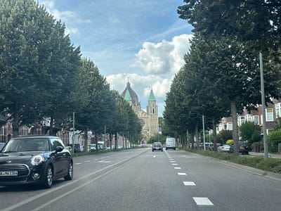The image shows a street in Maastricht, Netherlands, with a prominent church in the background. The street is lined with mature trees on both sides, creating a canopy over the road. A black Mini Cooper is in the foreground on the left, its license plate reading "GK MN 2304". Several other vehicles are visible on the road and parked along the side. The church, which appears to be a cathedral, features a large dome and spires. The sky is partly cloudy with some cumulus clouds. The architecture of the buildings along the street suggests a European city. The overall scene is one of a peaceful urban environment on a day with moderate weather.