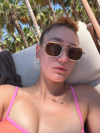 A woman is lying on a lounge chair under palm trees on a sunny day. She is wearing a pink bikini and sunglasses. Her hair is pulled up in a bun. The background is a blur of palm trees and blue sky. The location is identified as Keramoti, Greece.